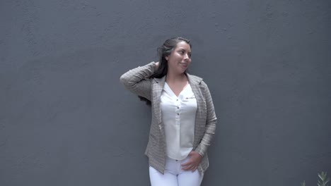 happy girl leaning against a gray wall, smiling while fixing her hair, displaying confidence and happiness in her appearance and demeanor