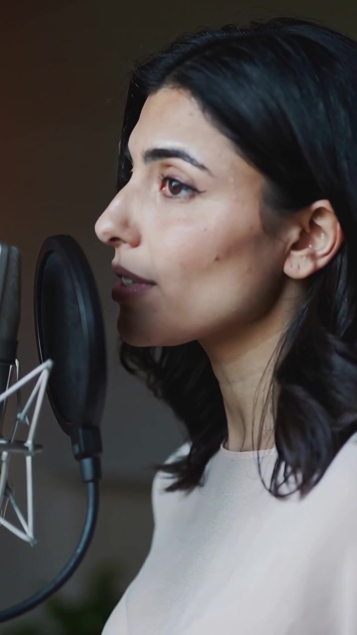 Video verticale: Segnale d'ascolto, cantante con top chiaro in uno studio casalingo, che canta in un microfono usando un filtro anti-pop.