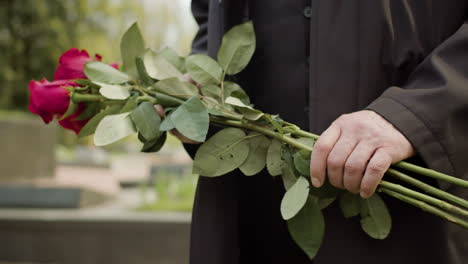 close up view of man in black raincoat and suit holding red roses and putting it on tombstone in a graveyard