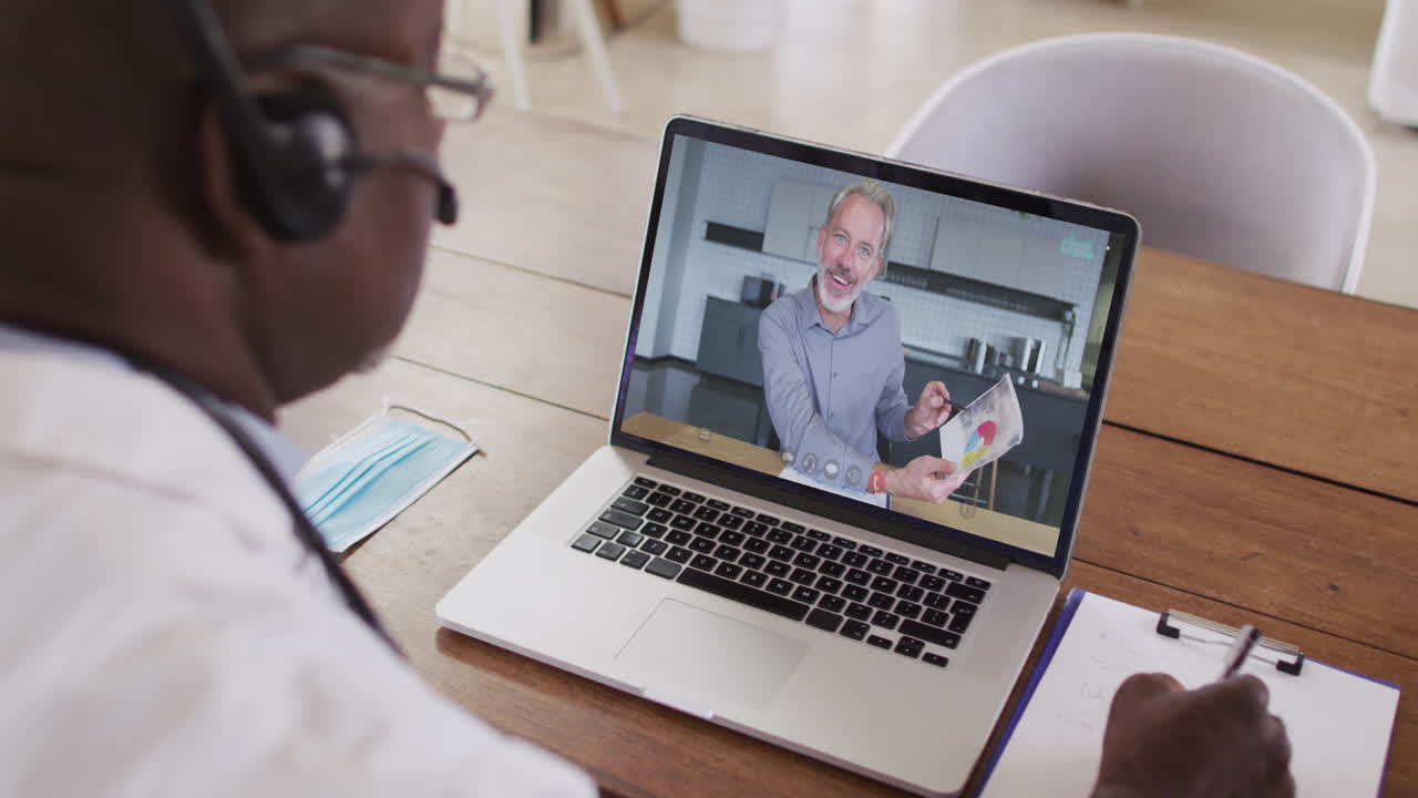 Ein afroamerikanischer Arzt mit Telefon-Headset macht sich Notizen während eines Videoanrufs am Laptop.