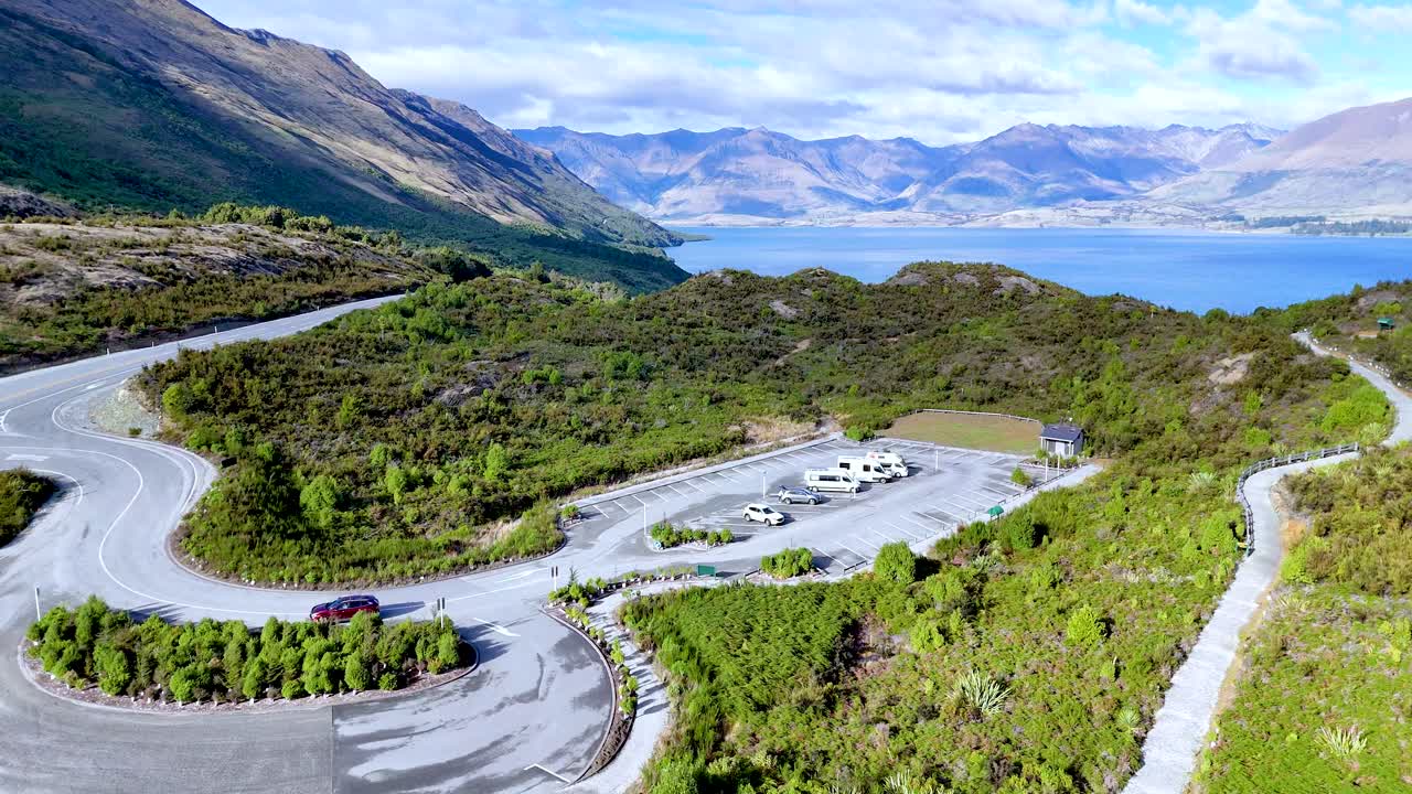 Drone glides above winding mountain road, parking lot, and scenic lake under bright daylight