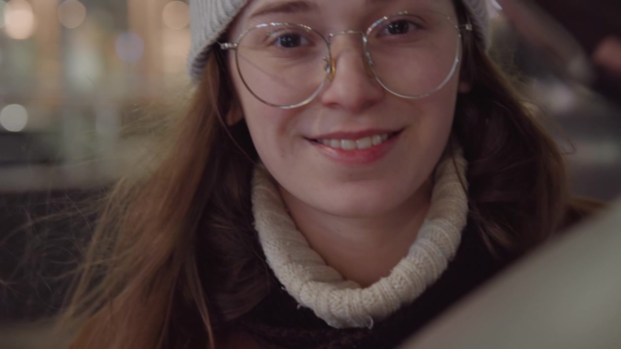 White woman student smiling at reflective sculpture, wearing beanie and glasses, turtleneck and coat, city lights bokeh, night plaza background, candid closeup capturing curious expression and soft