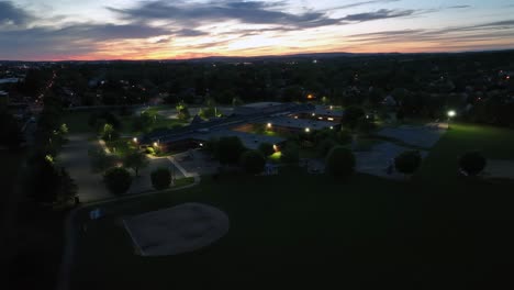 american school in warwick township during golden sunset. lighting facade of building at dusk. aerial approaching shot. empty parking area in evening.