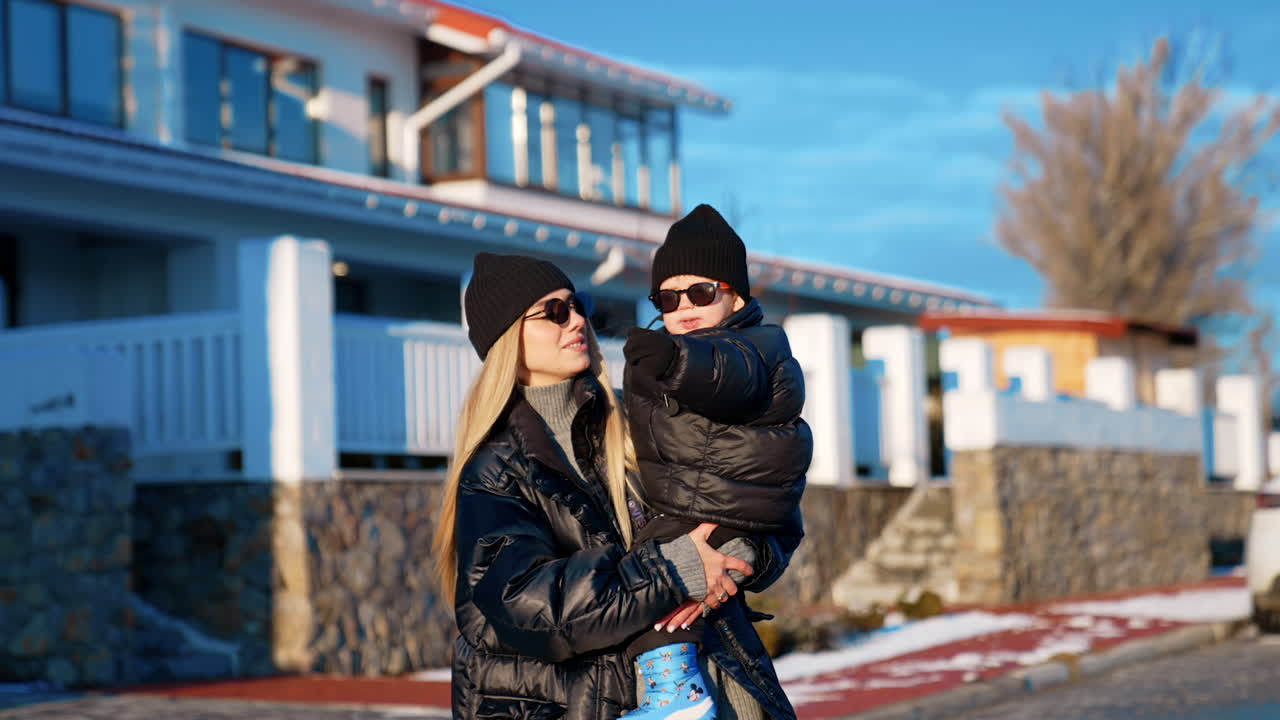 Mother and son wearing black caps, jackets and sunglasses outdoors. Happy woman turns around with her child.