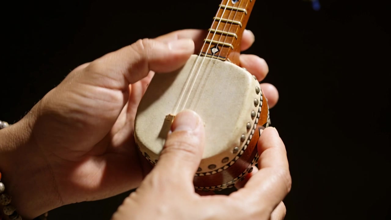 Le mani di un musicista entrano nell'inquadratura, pizzicando un mini banjo in uno studio, mostrando nappe e braccialetti di perline.