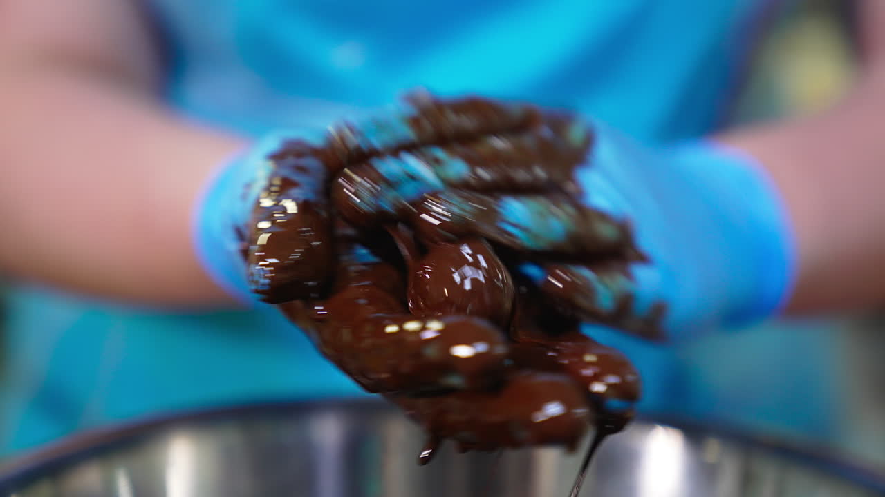 Gloved hand of confectioner smeared with black chocolate. Worker takes a candy and mixes it with chocolate. Close up.