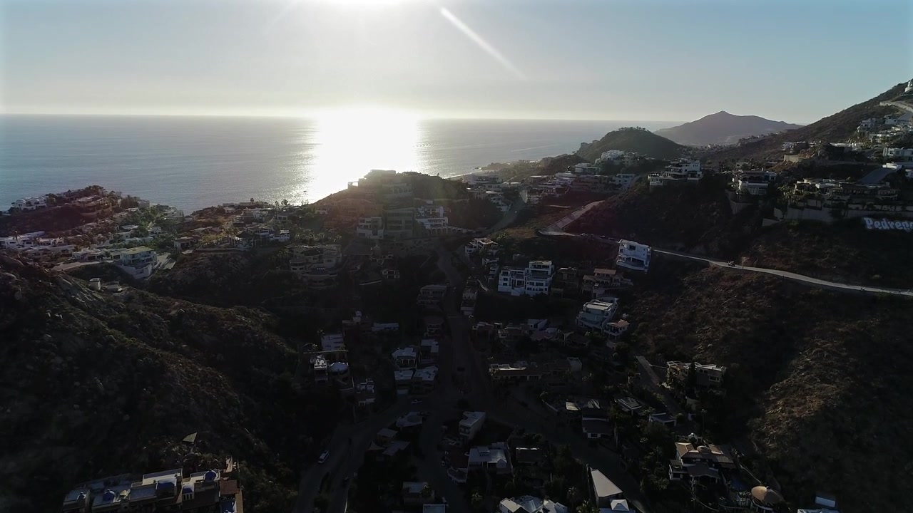 Un drone si innalza sulle colline di Pedregal a Cabo San Lucas, ricoperte di case verdi, rivelando l'Oceano Pacifico oltre, mentre la luce del sole attraversa l'obiettivo e scintilla sull'acqua