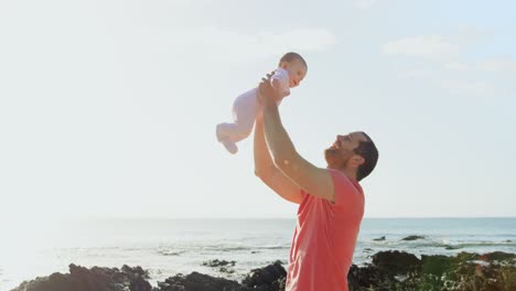 Side-view-of-mid-adult-caucasian-father-holding-baby-in-the-air-at-beach-on-a-sunny-day-4k