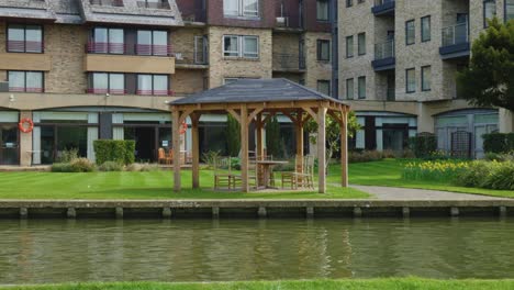 Pergola-next-to-calm-river-in-front-of-rustic-apartments-on-sunny-day