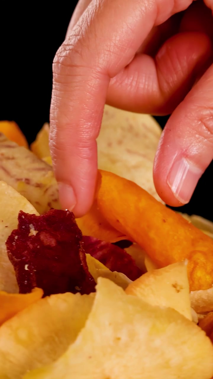 Una mano raccoglie chips croccanti di verdure miste a radice sotto l'illuminazione da studio, primo piano, sfondo nero, texture dettagliate