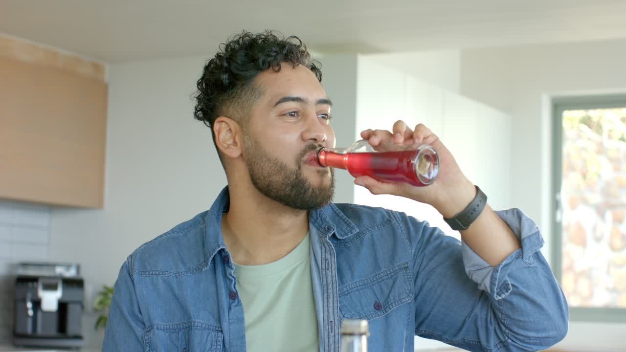 Mid adult man grabbing bottle at kitchen island, sipping and tasting flavor as friend enters