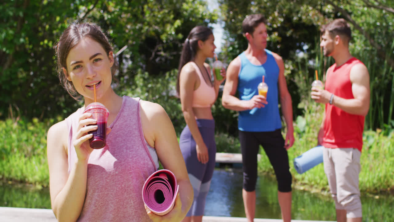 mujer caucásica sonriente bebiendo bebida saludable, con un grupo diverso hablando después de yoga en el parque soleado