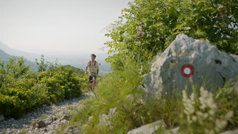 Hiker-walking-on-a-rocky-path-past-a-big-bush-and-a-rock-with-a-Knafelč-markation-for-hikers-indicating-the-right-path