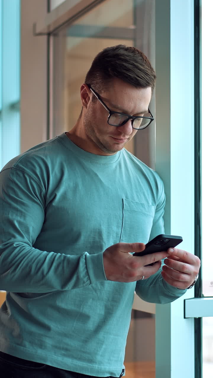 Fit strong dark-haired Caucasian man stands leaning on the window frame. Man in glasses focused on the phone he's holding in his hands. Vertical video