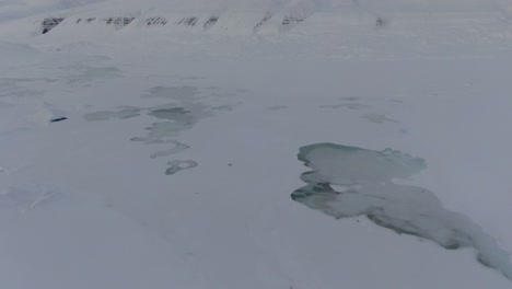 Drone-view-in-Svalbard-flying-over-snowy-white-mountains-with-frozen-lakes-in-Norway-on-a-cloudy-day