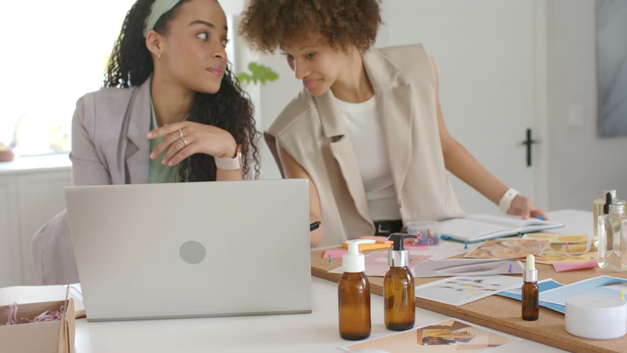 Due donne afroamericane si avvicinano, la fotocamera mostra dei campioni, mentre revisionano mockup di design su un laptop.