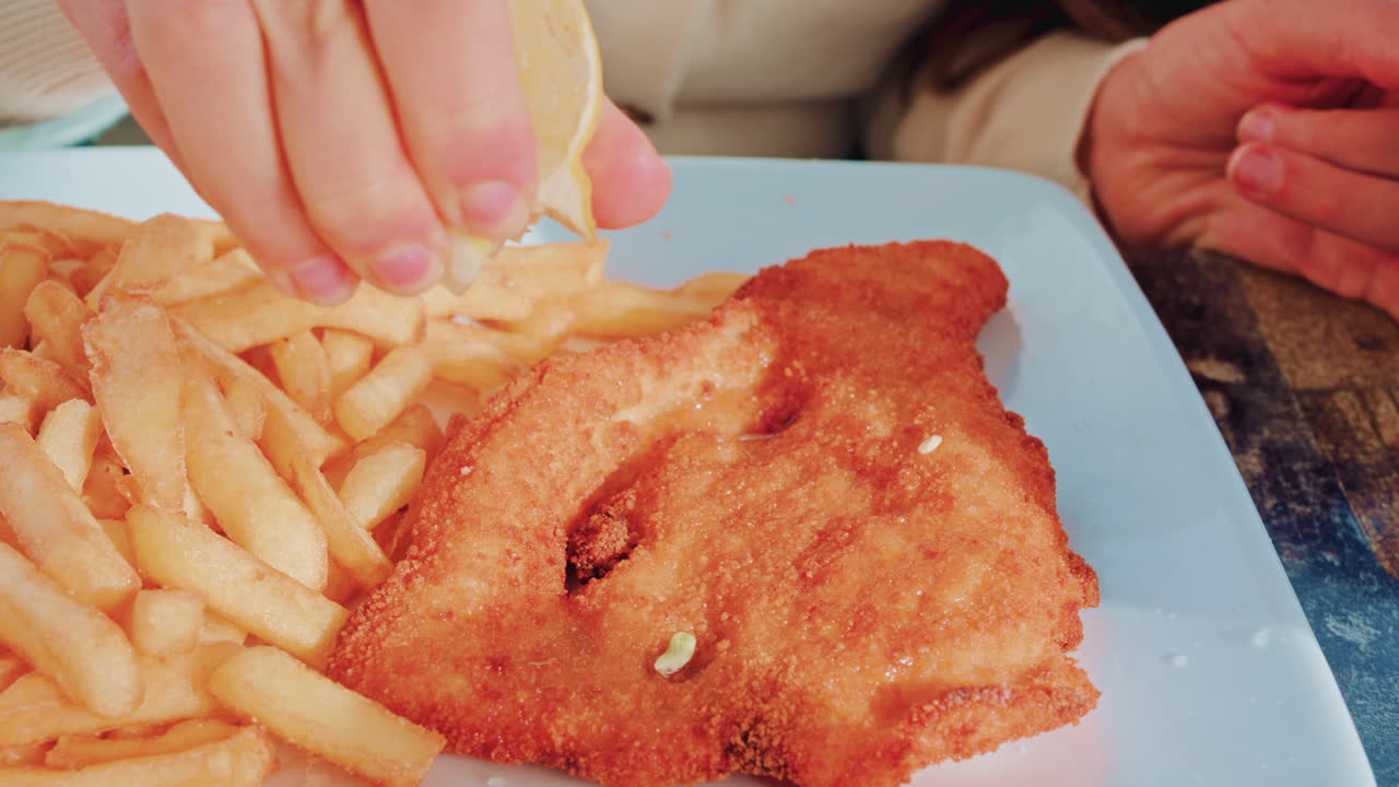 Crispy breaded fillet served with golden French fries on a blue plate as a hand sprinkles fresh lemon juice on top