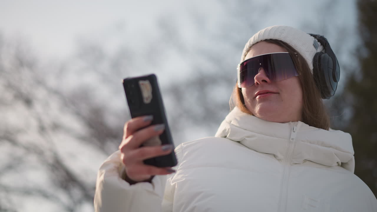 Young lady spinning with mobile in hands, body swaying under sunset light while scrolling screen on white winter jacket in snowy park setting, conveying chill tech vibe, casual modern outdoor moment