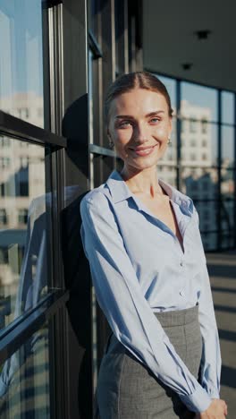 professional businesswoman leaning against large office window, radiating confidence while smiling and basking in natural sunlight, embodying corporate success and modern workplace elegance