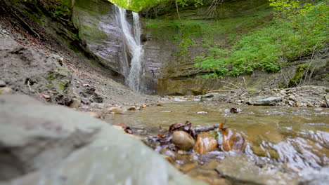 low angle shot of slowly flowing natural creek and waterfall in background surrounded by woodland during sunny day - slow motion pan shot