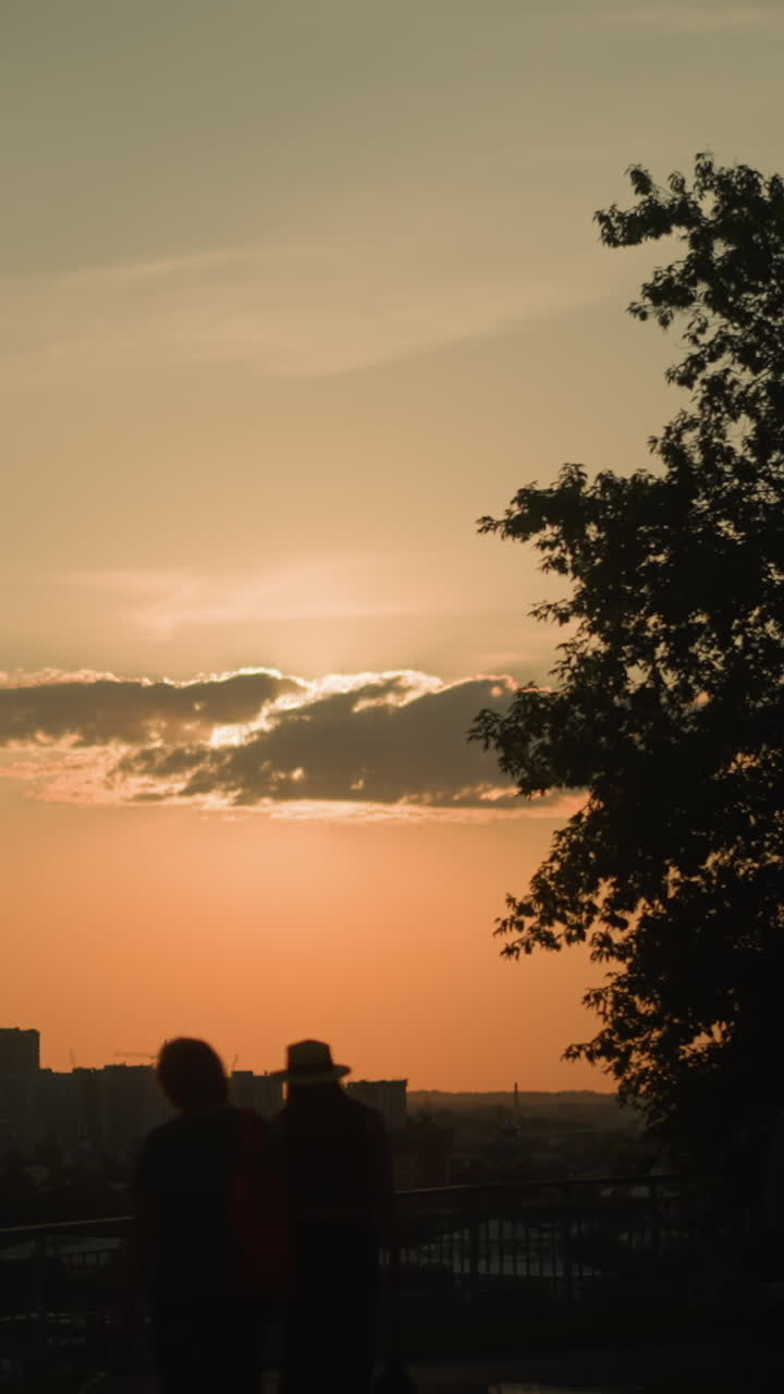 siluetas de personas caminando por el camino cerca de la barandilla de hierro al anochecer, con el paisaje urbano en el fondo, grandes árboles proyectando sombras, con la cálida luz dorada del sol iluminando el horizonte urbano