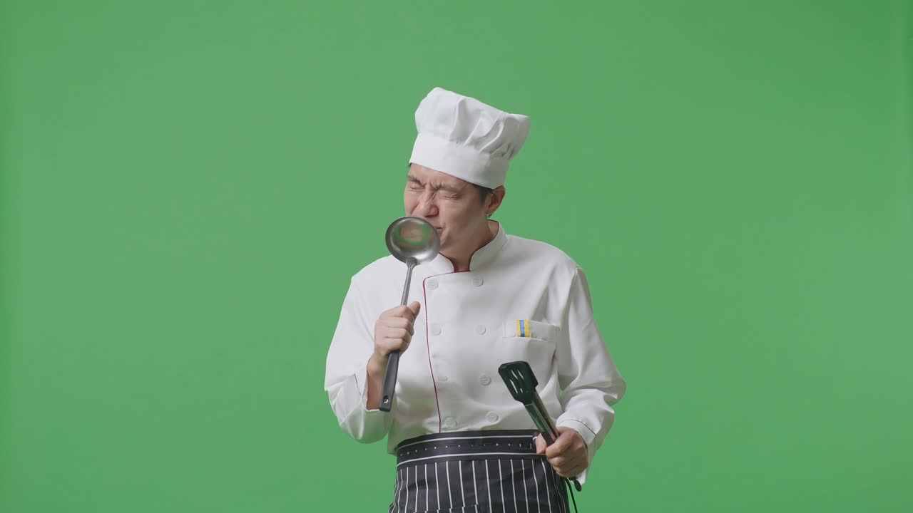 chef asiático con pinzas y cuchara y cantando mientras está de pie en el estudio de fondo de la pantalla verde