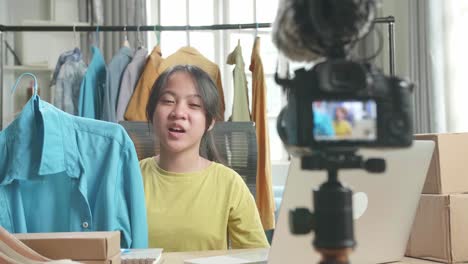 asian young girl online seller recommending a shirt and recording video by camera while using computer for selling clothes at home