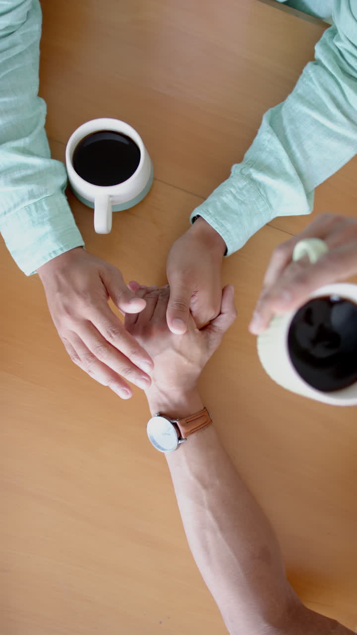 Vertical video of diverse gay male couple having coffee and holding hands at table, slow motion