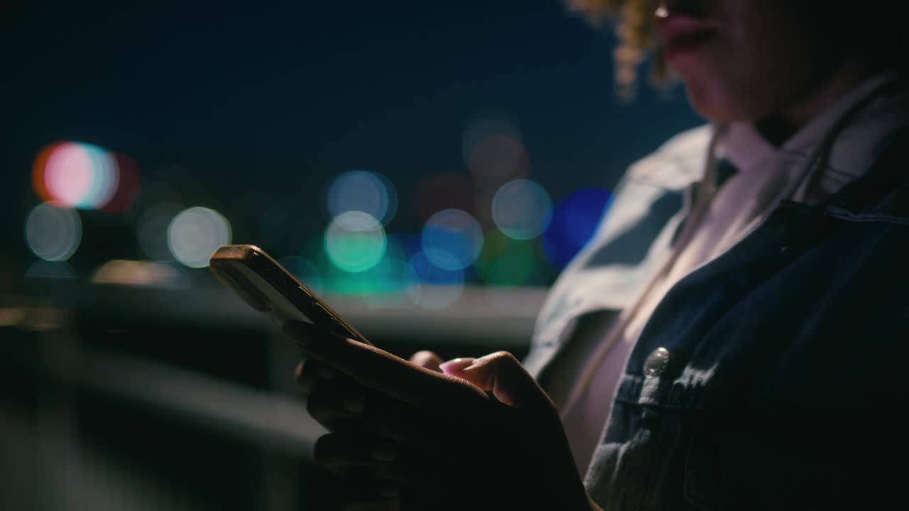 Detail of black woman standing on the bridge at night and browsing her mobile phone