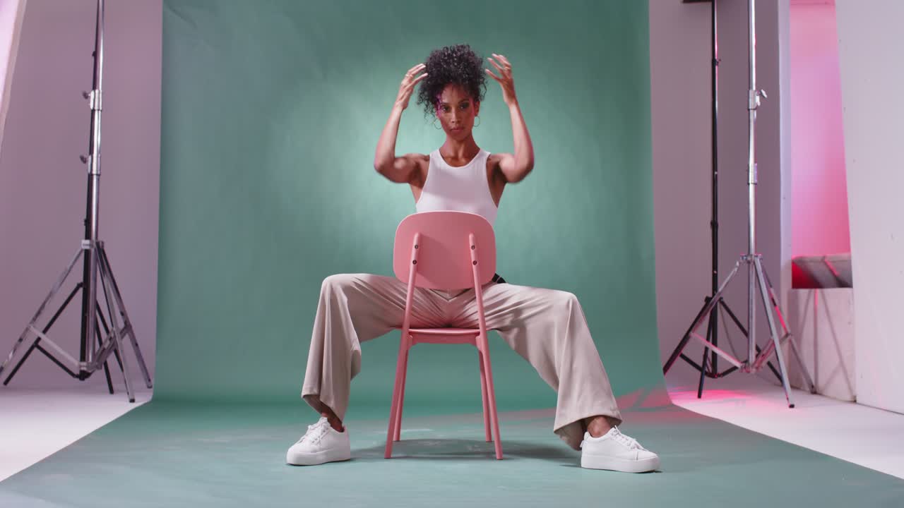 Donna afroamericana entra da destra, gira una sedia rosa e si esibisce in canottiera bianca in studio.