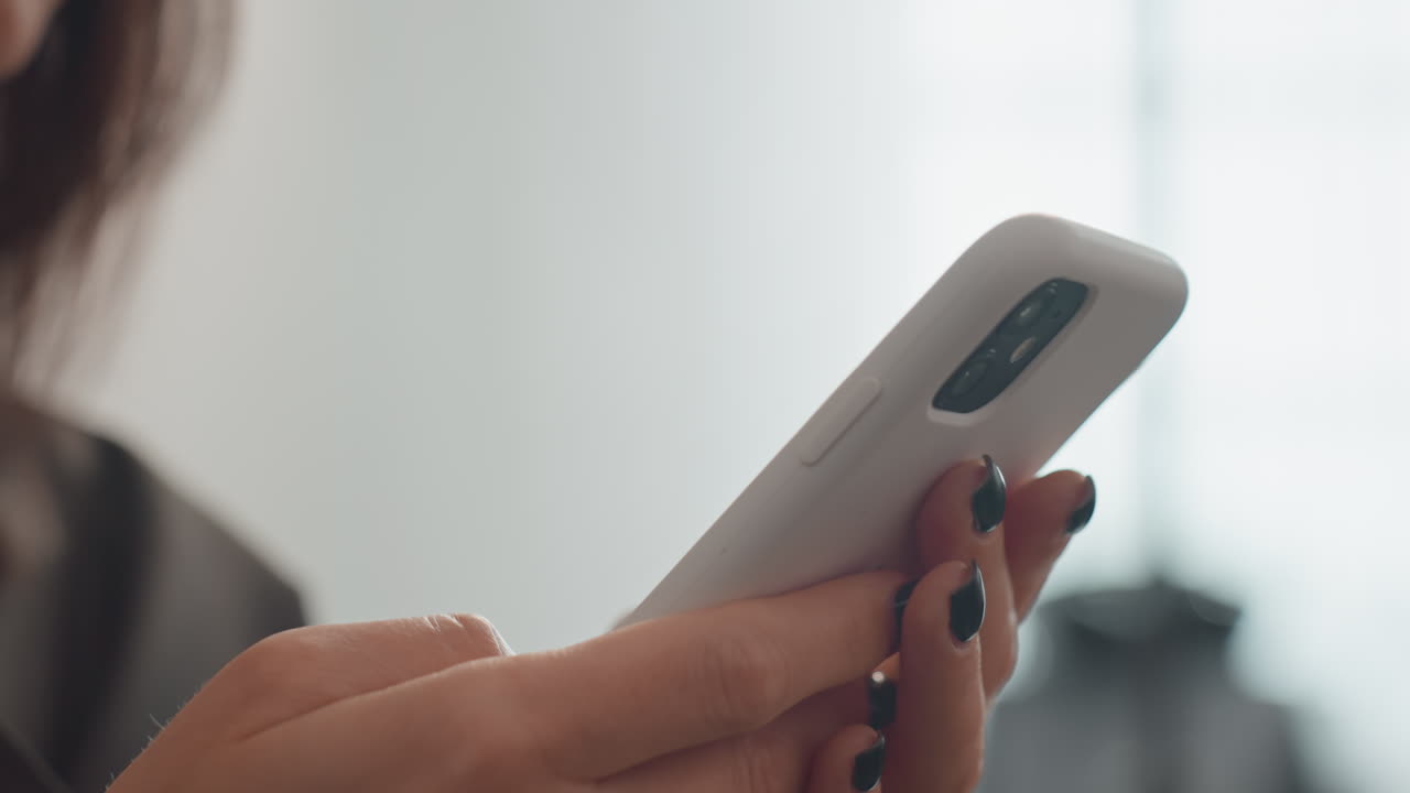 close das mãos de uma mulher jovem segurando um smartphone enquanto ela envia mensagens. Bokeh suave de contraluz, manicure escura, polegares deslizando mensagens em uma capa branca moderna, ambiente de escritório interno, clima calmo e íntimo, foco
