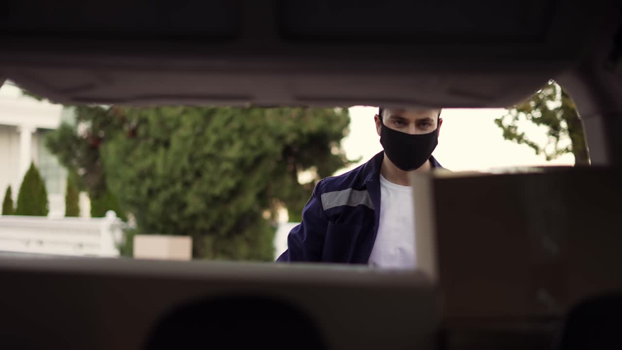 Delivery service. Delivery man in a protective mask and gloves opening the trunk and taking cardboard box parcel. Man closing