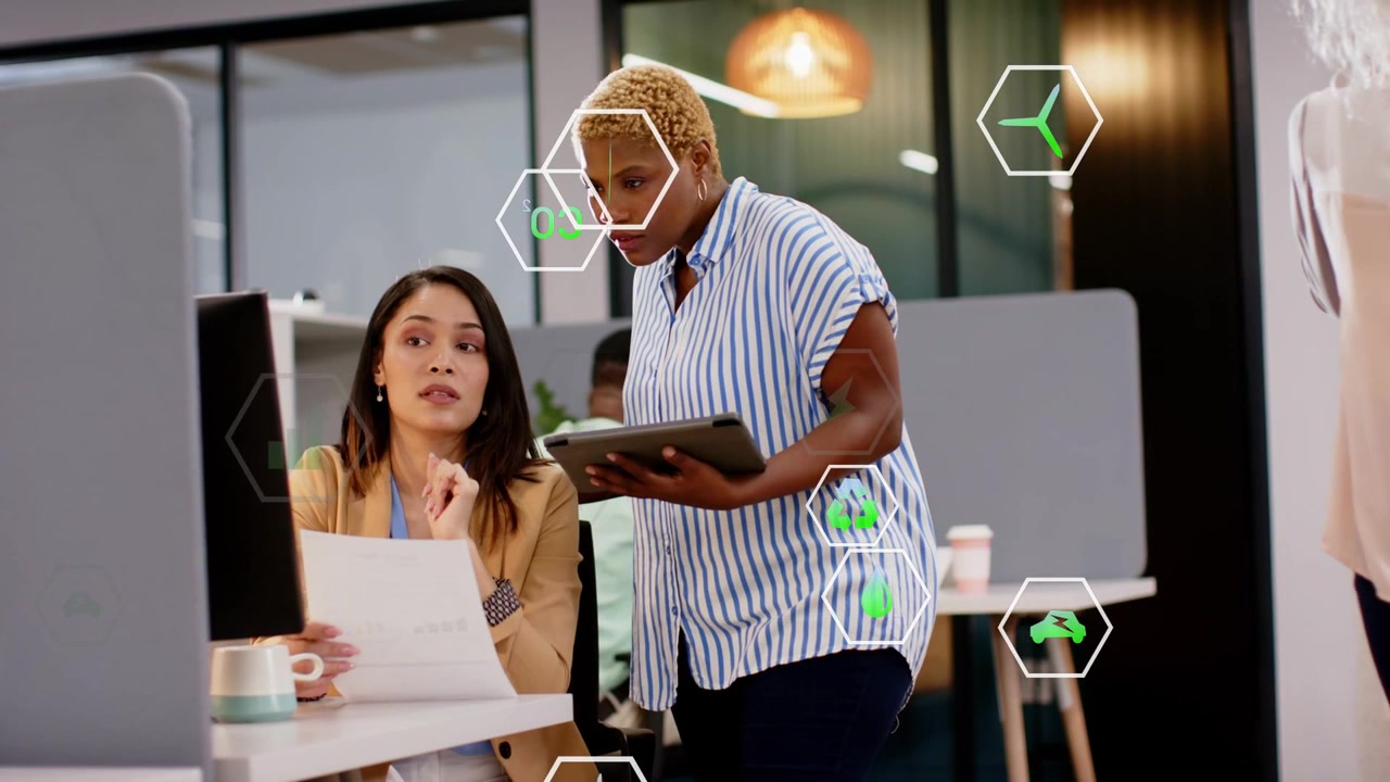 Femme assise gesticulant, un collègue penché, tous deux examinant un document, avec des icônes hexagonales de réalité augmentée flottant au-dessus d'eux dans un bureau.