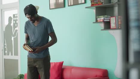 Young student wearing cap and casual clothing sits on red couch, drops one book beside him, crosses leg while holding another book, glancing down thoughtfully in cozy library