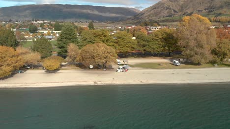 Dron-Aéreo-Que-Asciende-Desde-La-Playa-Con-Vistas-A-La-Ciudad-Y-Al-Lago-Wanaka,-Nueva-Zelanda-En-Otoño