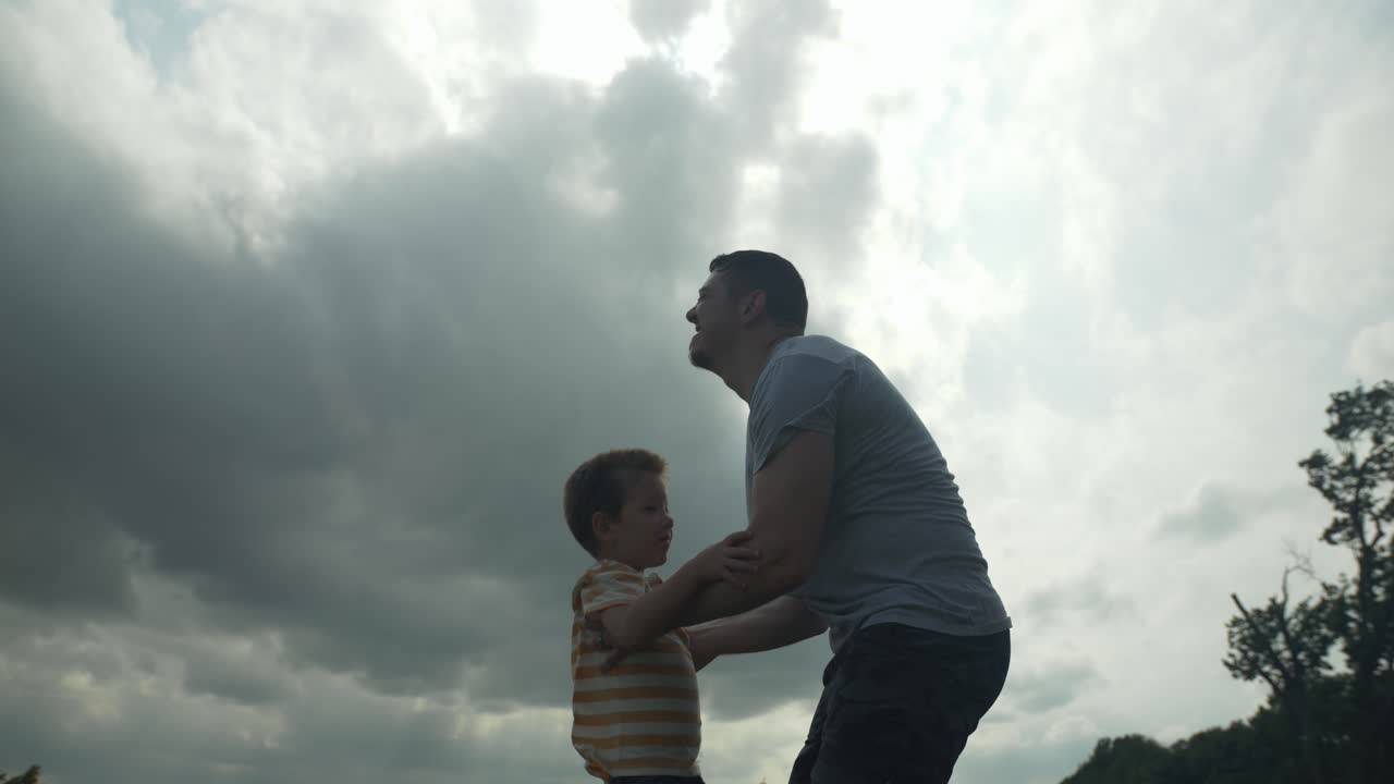 A happy, loving father holding and hugging his son for a warm, positive family moment outside under the sunlight shines through clouds.