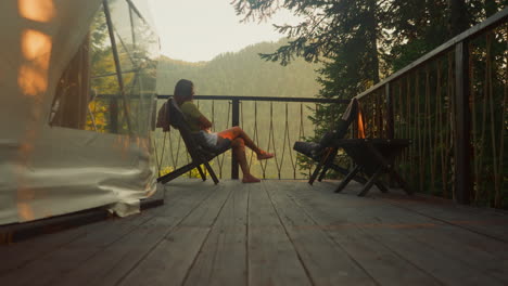 mujer solitaria descansa en un sillón en el balcón con vistas al bosque y las montañas con árboles al atardecer al anochecer temprano en la noche. paz y relajación en la soledad