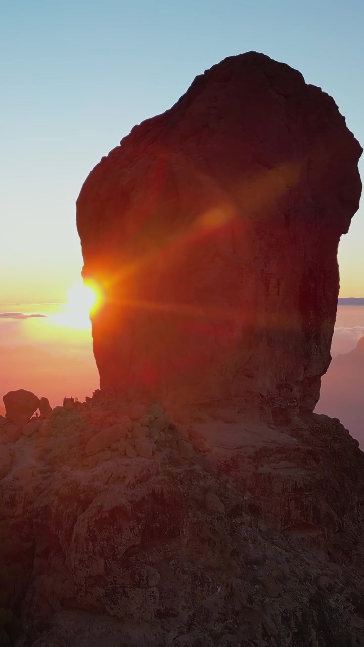 Stunning aerial view of the sun setting behind the iconic Roque Nublo, creating a dramatic silhouette and a vibrant lens flare over the clouds