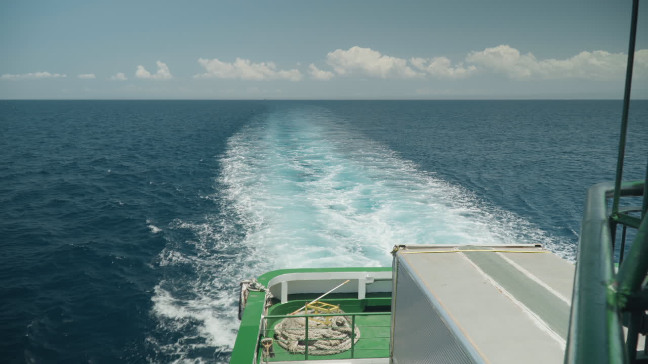 Heckwelle eines Bootes auf offenem Ozean, grünes Deck und Laderampe. Eine schaumige türkisfarbene Spur erstreckt sich bis zum fernen Horizont unter verstreuten Wolken. Ruhiges blaues Wasser, Mittagslicht, sanfter Wellengang, der Langsamkeit vermittelt.