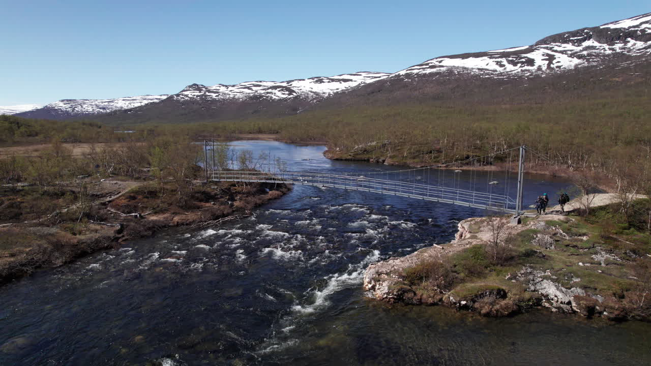 Three Hikers Walking over a Suspension Bridge on the Kungsleden near Abiskojaure in The Swedish Mountains, Backpacking in the Forest, Drone Pushing in