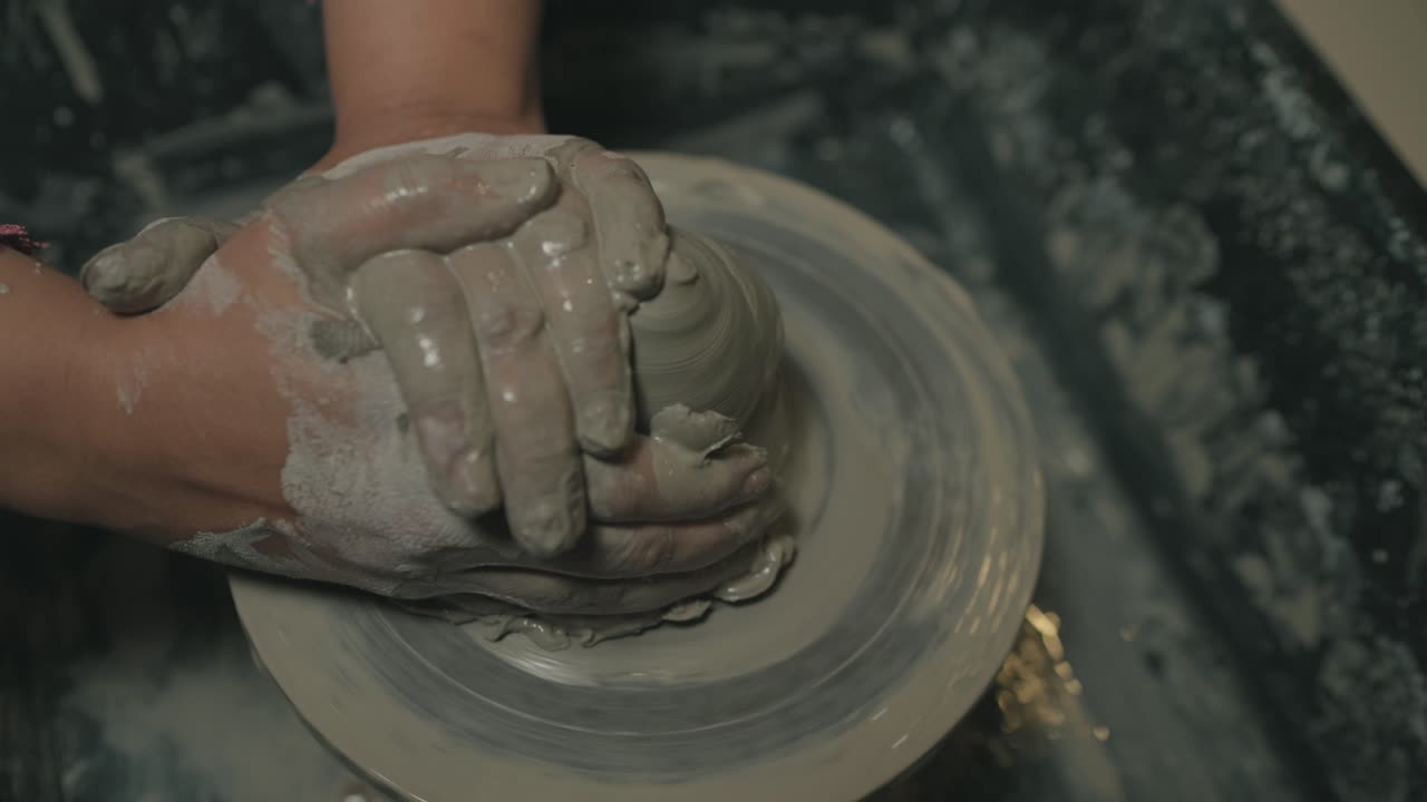 Close-up captures artisan's hands shaping wet clay on spinning pottery wheel, fingers covered in mud while molding the form under workshop lighting, showcasing delicate ceramic creation process