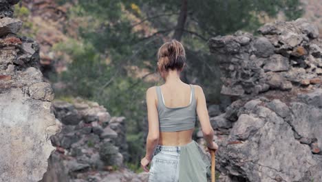 Rear-view-woman-with-wooden-stick-walks-by-rocky-hills-or-ruins,-close-up