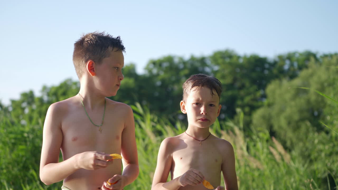 Hungry boys eating on the natural green background. Two brothers with bare bodies standing outdoors and eat tasty food in summer. Happy childhood.