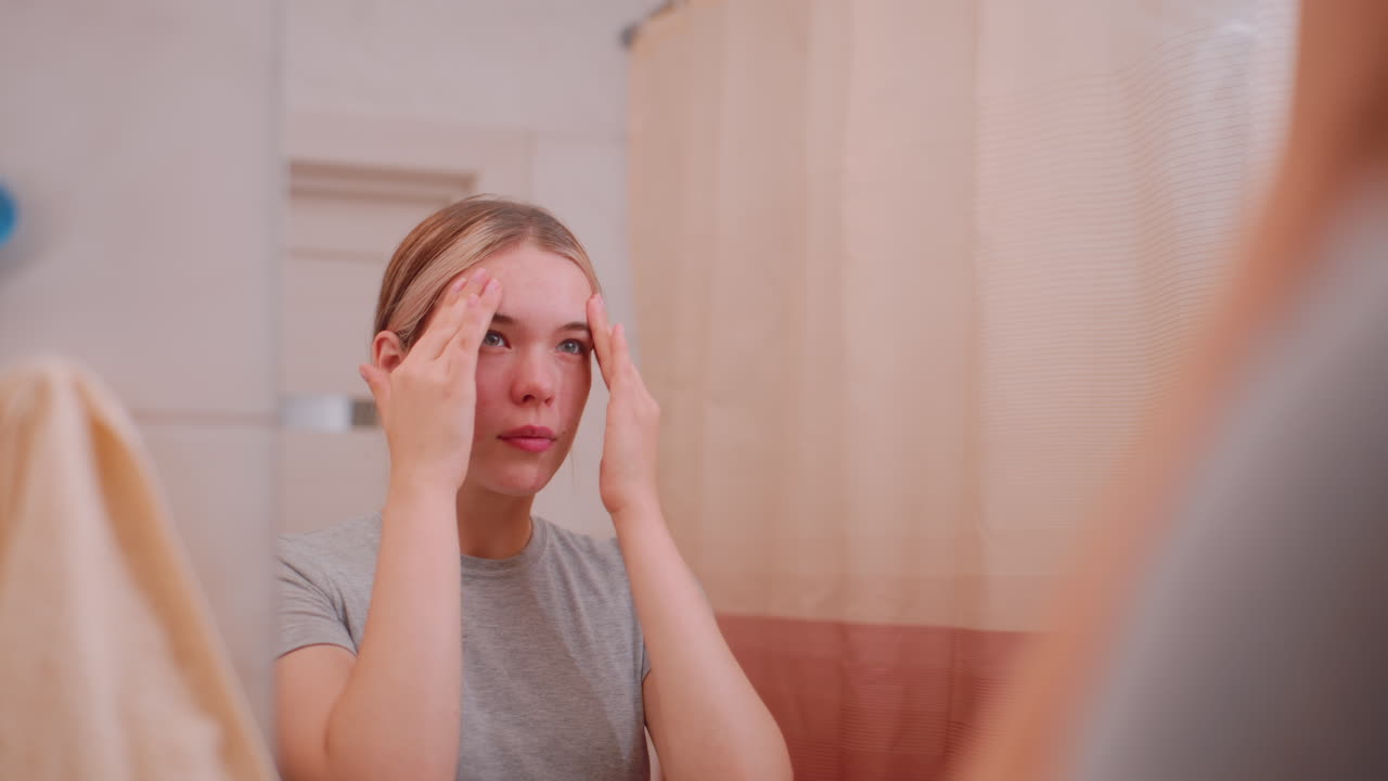 Teenager applying lotion on face while standing in bathroom mirror, focusing on skincare, personal grooming, wellness, beauty care, and self care promoting clean healthy skin