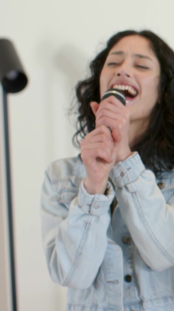 Vertical video: Closing eyes, woman singing emotively in studio with mic, denim jacket, mic stand