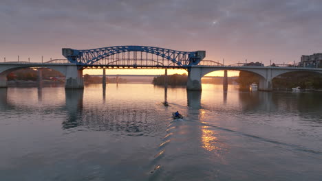 aerial footage during sunrise close to the water of the tennessee river following rowers on a boat going under the market street bridge during sunrise