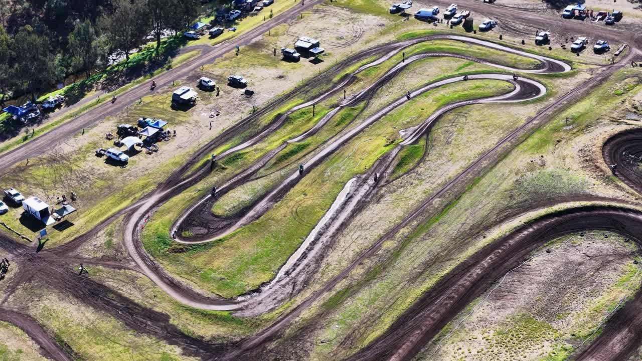 Motocross riders speed around winding dirt track, aerial perspective, bright daylight, dynamic movement