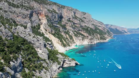 Costa-De-Baunei,-Playa-De-Cala-Goloritze-Y-Barcos-Turísticos-En-El-Mar-Azul-Turquesa-En-Cerdeña,-Italia---Antena-4k