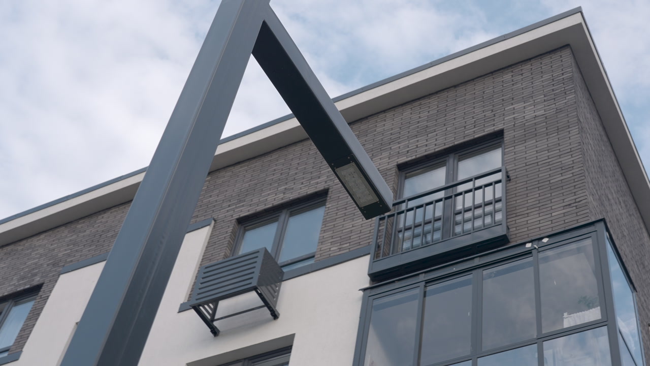 Angular Lamppost Arm Against Cloudy Sky, Closeup Of Metal Bracket Beside Brick Apartment Facade With Balcony
