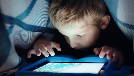 happy little boy using digital tablet computer under blanket enjoying learning on touchscreen technology playing games having fun at bedtime